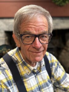 Walt Woodward, wearing glasses, a plaid shirt, and suspenders, from the chest up. He is smiling and in front of a fireplace.